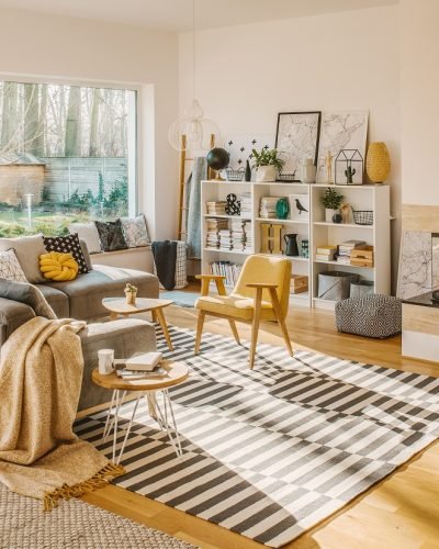 Blanket thrown on grey corner sofa in white living room interior with fresh plants, carpet, fireplace and simple posters