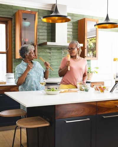 Senior African American woman and senior biracial woman are cooking together in a modern kitchen. They share a joyful moment, tasting food and bonding over the culinary experience.