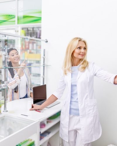 smiling female pharmacist taking pills from shelf while customer pointing with finger